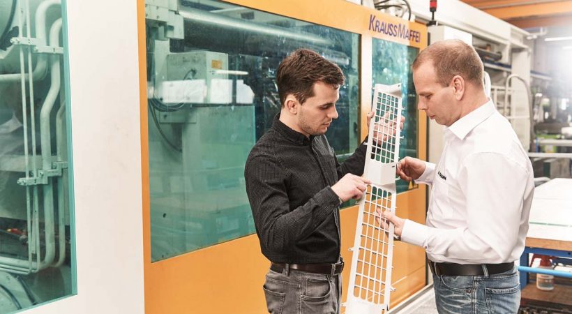 Zwei Kollegen besprechen ein Kunststoffteil vor einer Spritzguss-Maschine von Krauss Maffei